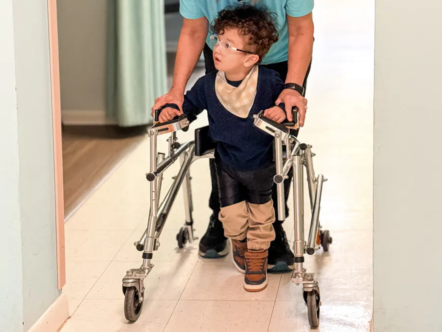 A male child using a walker with a therapist to walk in a hallway